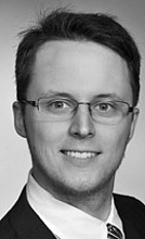 Alexander Groll Schwarzweiß-Porträt eines Mannes mit Brille, der ein formelles Outfit trägt. Er lächelt freundlich in die Kamera, während der Hintergrund neutral gehalten ist. Die Bildkomposition vermittelt ein professionelles und selbstbewusstes Auftreten.